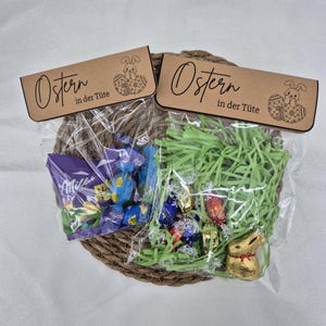 Op de afbeelding: Twee paasgeschenktassen gevuld met snoep en groen papiergras, gepresenteerd in een gevlochten mand. Elke tas heeft een bruin label met het Duitse woord "Ostern" en een konijnenillustratie.