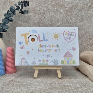 Peut inclure: Un panneau rectangulaire blanc sur un petit chevalet en bois avec le texte TOLL dass du mich begleitet hast! et Danke für alles! en lettres colorées. Le design inclut des illustrations d'un soleil, d'une maison, de fleurs et d'un arrosoir.