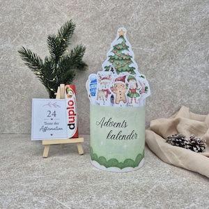 Puede incluir: Un recipiente cilíndrico Advents kalender con base verde y un adorno de árbol de Navidad con ilustraciones de un reno, un hombre de jengibre y un elfo. Al lado, un caballete de madera sostiene una tarjeta 24 Texte der Affirmation.