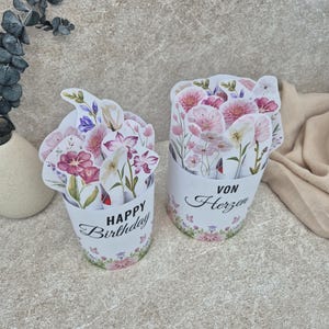 May include: Two cylindrical paper containers filled with die-cut floral illustrations. The left container features the text "HAPPY Birthday" in black script and block letters. The right container displays the German text "VON Herzen" in black.