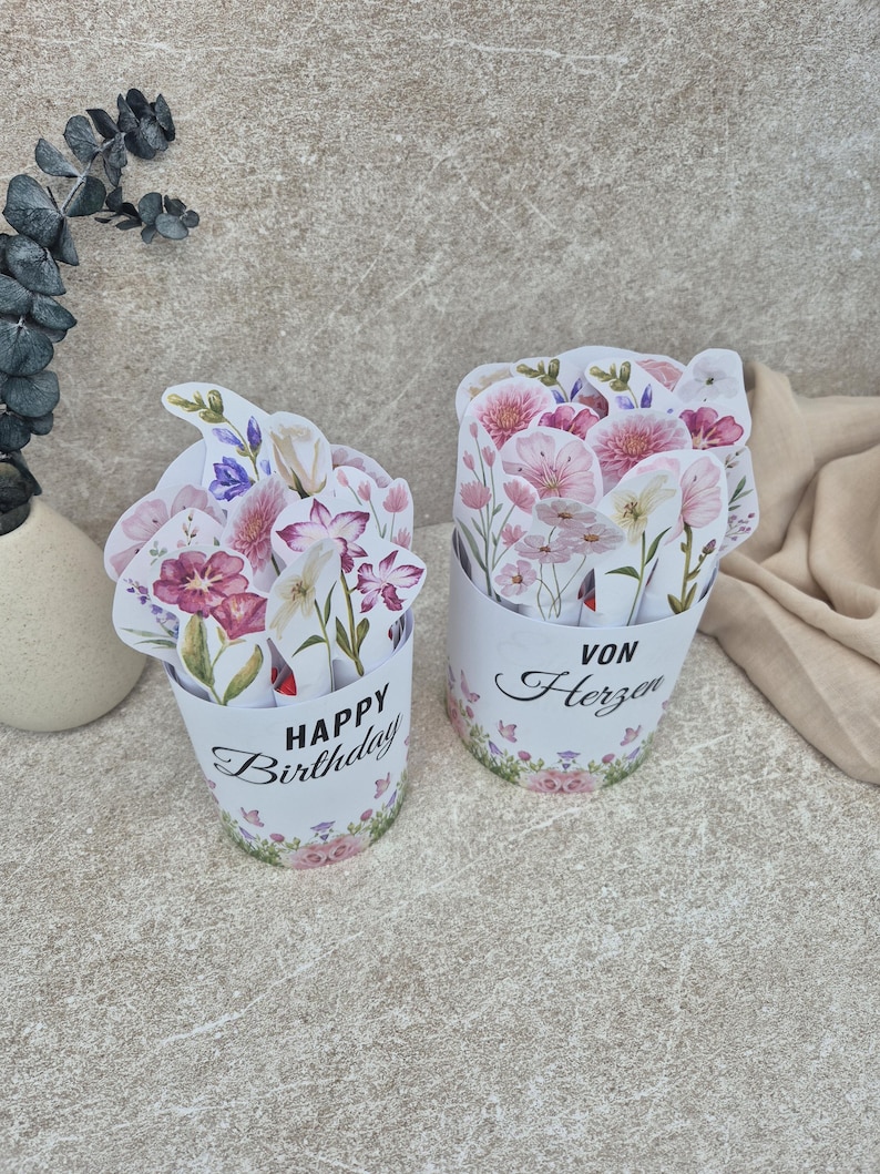 May include: Two cylindrical paper containers filled with die-cut floral illustrations. The left container features the text "HAPPY Birthday" in black script and block letters. The right container displays the German text "VON Herzen" in black.