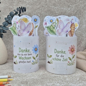 Könnte beinhalten: Zwei weiße zylindrische Tassen mit bunten Illustrationen von Raupen, Schmetterlingen und Blumen. Die linke Tasse trägt die Aufschrift "Danke, dass du mir beim Wachsen geholfen hast", die rechte "Danke, für die schöne Zeit". Beide Tassen haben dekorative Aufsätze.