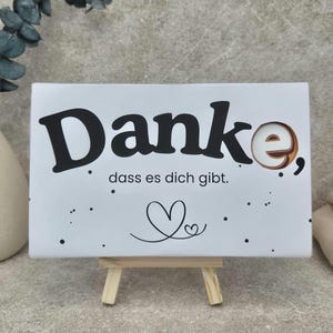 Op de afbeelding: Een rechthoekig wit bordje op een kleine houten ezel. Het bordje bevat de zwarte tekst "Danke, dass es dich gibt." en een kleine lijntekening van een hartje. De letter "e" in "Danke" is gestileerd met een bruin en wit cirkelvormig ontwerp.