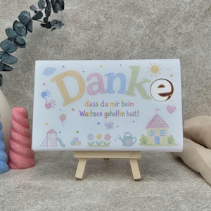 May include: A rectangular white sign on a small wooden easel featuring the text "Danke dass du mir beim Wachsen geholfen hast!" in colorful crayon-style lettering. The design includes illustrations of a house, flowers, a watering can, and a playground.
