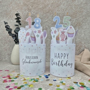 Op de afbeelding: Twee cilindervormige witte houders met kleurrijke stippen. De linkerhouder bevat de tekst "Herzlichen Glückwunsch" en een taarttopper met het getal 18. De rechterhouder bevat de tekst "HAPPY Birthday" en een topper met het getal 25.