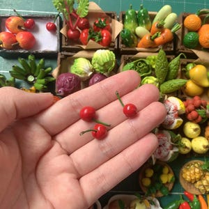 May include: A hand holding three miniature red cherries with green stems. The cherries are surrounded by a variety of miniature fruits and vegetables, including bananas, cucumbers, lettuce, and peppers.