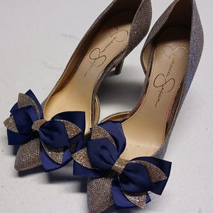 May include: A pair of Jessica Simpson high heels with a glittery silver finish. Each shoe features a large navy blue bow with gold glitter accents. The heels have a pointed toe and a low heel.