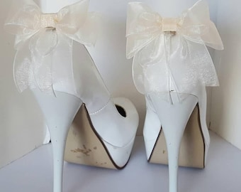 May include: A pair of white high-heeled shoes with a platform sole. Each shoe has a large ivory bow on the back.