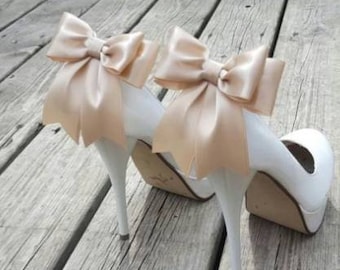 May include: A pair of white high-heeled shoes with platform soles. Each shoe features a large, beige satin bow at the heel. The heels are slender and pointed. The shoes are displayed on a wooden surface.
