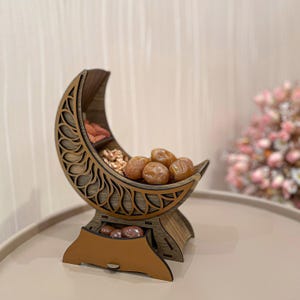 May include: Wooden crescent-shaped serving dish with intricate cut-out designs. The dish holds dates, walnuts, and other items. The base has a small drawer. The color is a warm brown.