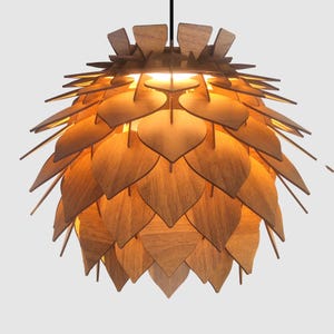 Op de afbeelding: Een houten hanglamp met een gelaagd ontwerp. De lamp is gemaakt van vele houten, bladvormige stukken, wat een unieke, natuurlijke uitstraling creëert. De armatuur hangt aan een zwart snoer en straalt een warme gloed uit.