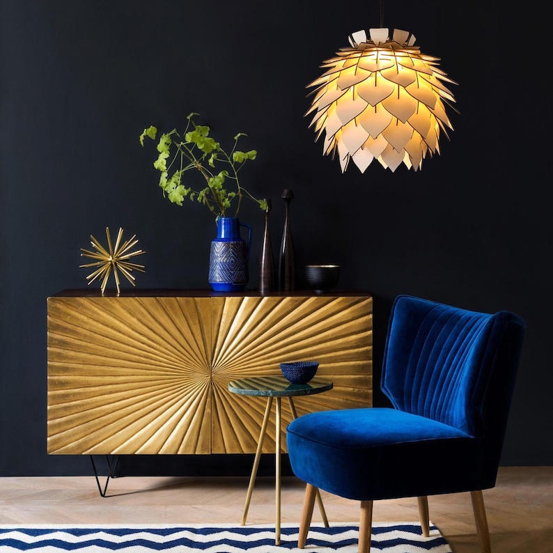 May include: A gold-colored cabinet with a sunburst design is the focal point of this interior shot. A blue velvet chair and a small side table with a blue bowl are in the foreground. A decorative light fixture hangs from the ceiling.
