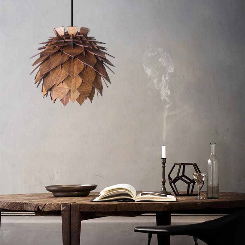 May include: A wooden pendant light fixture with a layered, leaf-like design hangs above a rustic wooden table. The warm brown tones of the light contrast with the neutral background. A candle burns, and a book is open on the table.