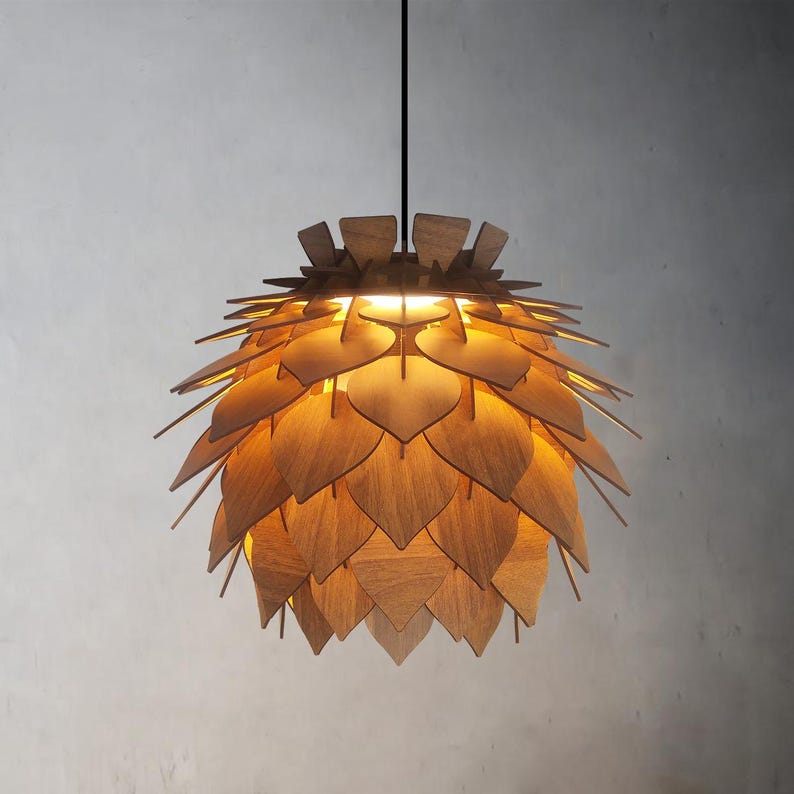 May include: A wooden pendant light fixture with a layered, leaf-like design. The light is suspended from a black cord and emits a warm glow. The fixture is made of light-colored wood and has a unique, natural aesthetic.