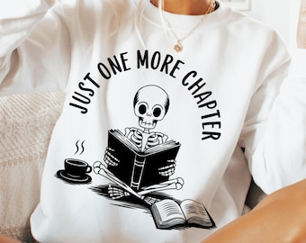 May include: White sweatshirt with a black skeleton reading a book. The text on the sweatshirt says "Just One More Chapter".