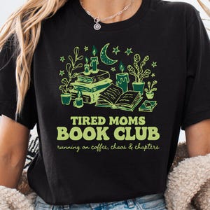 May include: Black t-shirt with a green graphic design featuring books, candles, and plants. The text reads "TIRED MOMS BOOK CLUB" with the tagline "running on coffee, chaos & chapters". The design includes a crescent moon and stars.