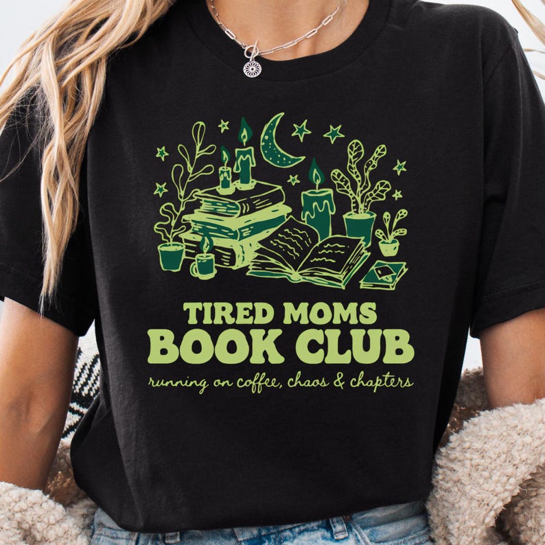 May include: Black t-shirt with a green graphic design featuring books, candles, and plants. The text reads "TIRED MOMS BOOK CLUB" with the tagline "running on coffee, chaos & chapters". The design includes a crescent moon and stars.