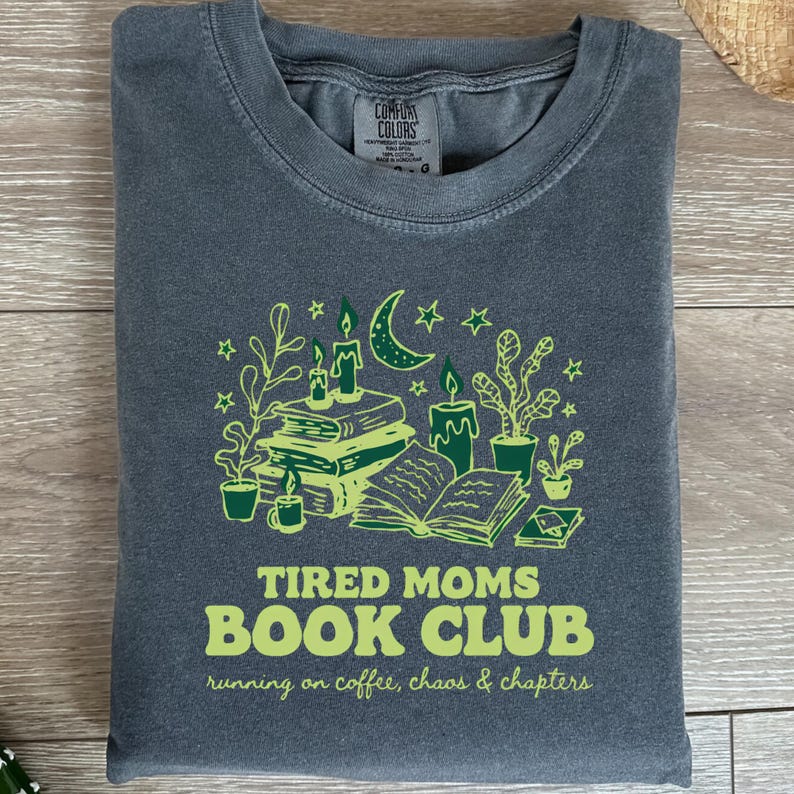 May include: A folded charcoal gray t-shirt with a green graphic. The graphic features books, candles, plants, a crescent moon, and stars. The text reads "Tired Moms Book Club" and "running on coffee, chaos & chapters."