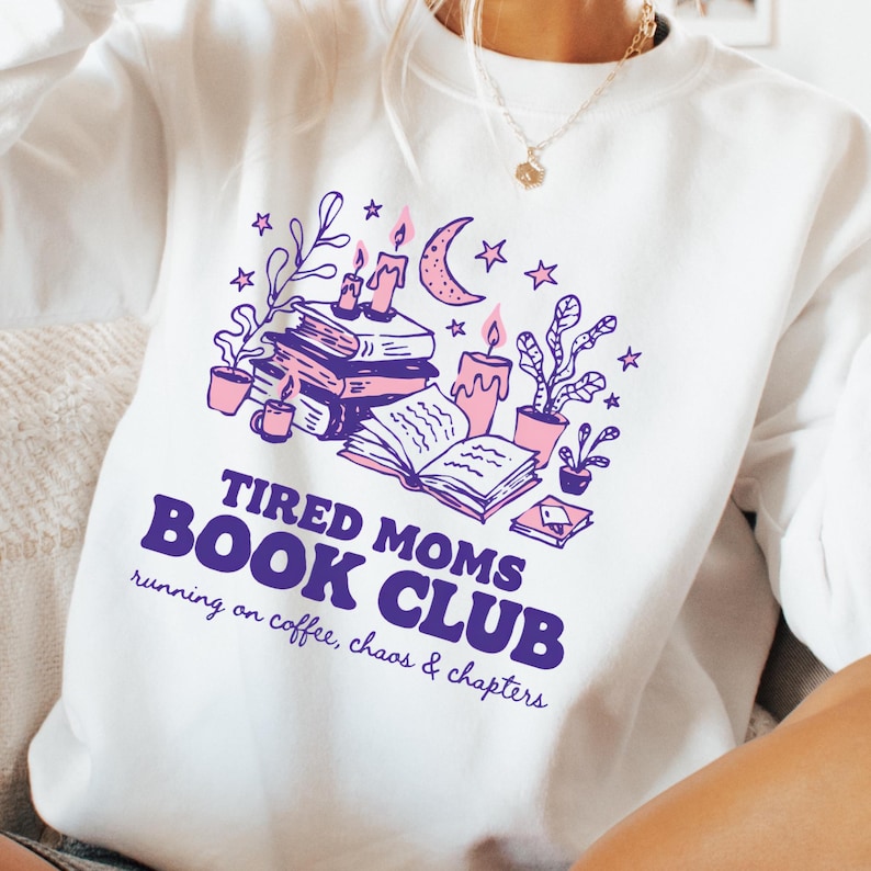May include: White sweatshirt with a purple and pink graphic. The graphic includes books, candles, plants, stars, and the text "TIRED MOMS BOOK CLUB running on coffee, chaos & chapters."