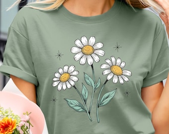 以下が含まれることがあります： セージグリーンのTシャツ。デイジーの花柄のデザイン。グラフィックは、黄色の中心と緑の茎と葉を持つ3つの白いデイジーを示しています。デザインは小さなスターバーストでアクセントされています。