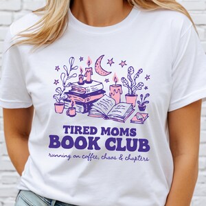 May include: White t-shirt with a purple and pink graphic. The graphic includes books, candles, plants, and a crescent moon. Text reads "TIRED MOMS BOOK CLUB" with "running on coffee, chaos & chapters" below.