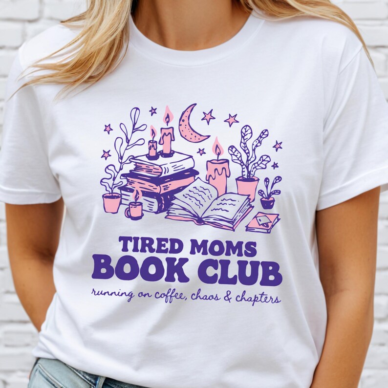 May include: White t-shirt with a purple and pink graphic. The graphic includes books, candles, plants, and a crescent moon. Text reads "TIRED MOMS BOOK CLUB" with "running on coffee, chaos & chapters" below.
