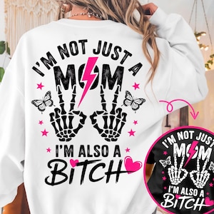 以下が含まれることがあります： 「I'M NOT JUST A MOM I'M ALSO A BITCH」の文字が入った白いスウェットシャツ。デザインには、ピンクの稲妻、ピースサインの骸骨の手、蝶、ピンクの星が含まれています。デザインの小さなバージョンが右下にあります。