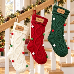 May include: Three cable-knit Christmas stockings in white, red, and green, each featuring a ribbed cuff, a leather-like name tag engraved with the name Amelia, and small decorative pom-poms in contrasting colours attached to the front.