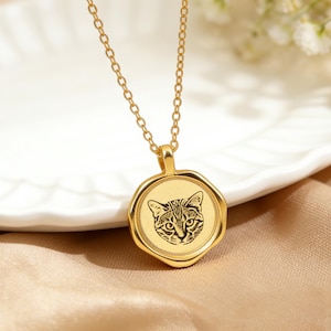 Può includere: Collana color oro con un ciondolo raffigurante la faccia di un gatto. Il ciondolo è rotondo con un bordo decorativo e una catena. Il volto del gatto è un'illustrazione dettagliata in bianco e nero.