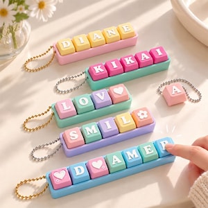 Può includere: Una collezione di portachiavi rettangolari con tasti meccanici colorati che presentano lettere, cuori e icone di fiori. I tasti sono montati su basi di plastica in tonalità pastello rosa, turchese, viola e blu con catenelle a palline in metallo.