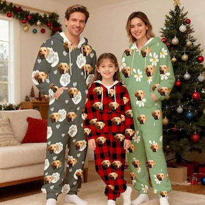 Può includere: Tre persone che indossano pigiami con cappuccio coordinati. L'adulto a sinistra indossa un pigiama grigio con impronte di zampe e facce di cani. Il bambino indossa un pigiama a quadri rossi con facce di cani. L'adulto a destra indossa un pigiama verde con margherite e facce di cani.