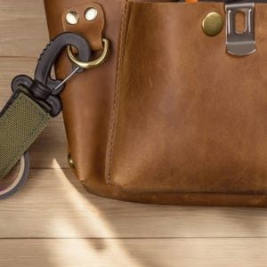 Pu&ograve; includere: Una borsa porta attrezzi in pelle marrone con un moschettone verde e nero attaccato a un rotolo di nastro adesivo. La borsa ha una fibbia in ottone e una chiusura in metallo.