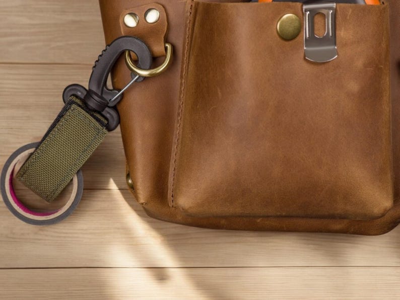 Pu&ograve; includere: Una borsa porta attrezzi in pelle marrone con un moschettone verde e nero attaccato a un rotolo di nastro adesivo. La borsa ha una fibbia in ottone e una chiusura in metallo.