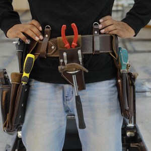 Può includere: Una cintura portautensili in pelle marrone con più tasche e passanti. La cintura è indossata da una persona ed è piena di vari strumenti, tra cui un martello, una pinza e un cacciavite.