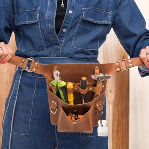 Pu&ograve; includere: Una cintura portautensili in pelle marrone con pi&ugrave; tasche e una variet&agrave; di strumenti, tra cui un martello, pinze e cacciaviti. La cintura &egrave; indossata da una persona che indossa una tuta di jeans blu.