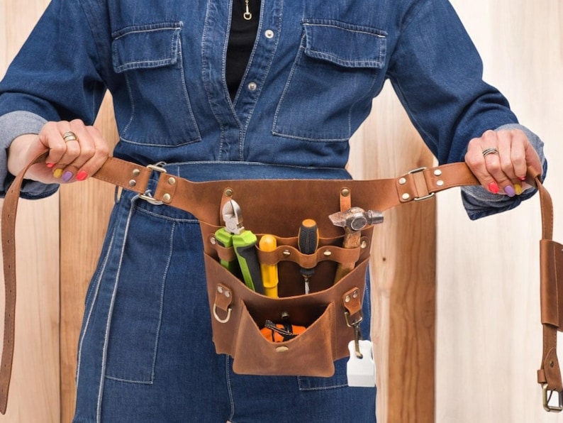 Pu&ograve; includere: Una cintura portautensili in pelle marrone con pi&ugrave; tasche e una variet&agrave; di strumenti, tra cui un martello, pinze e cacciaviti. La cintura &egrave; indossata da una persona che indossa una tuta di jeans blu.