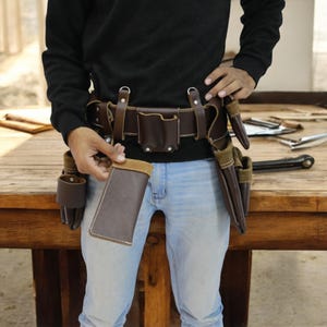 May include: A brown leather tool belt with multiple pockets and pouches. The belt is worn by a person and is secured with a metal buckle. The belt is on a wooden surface.