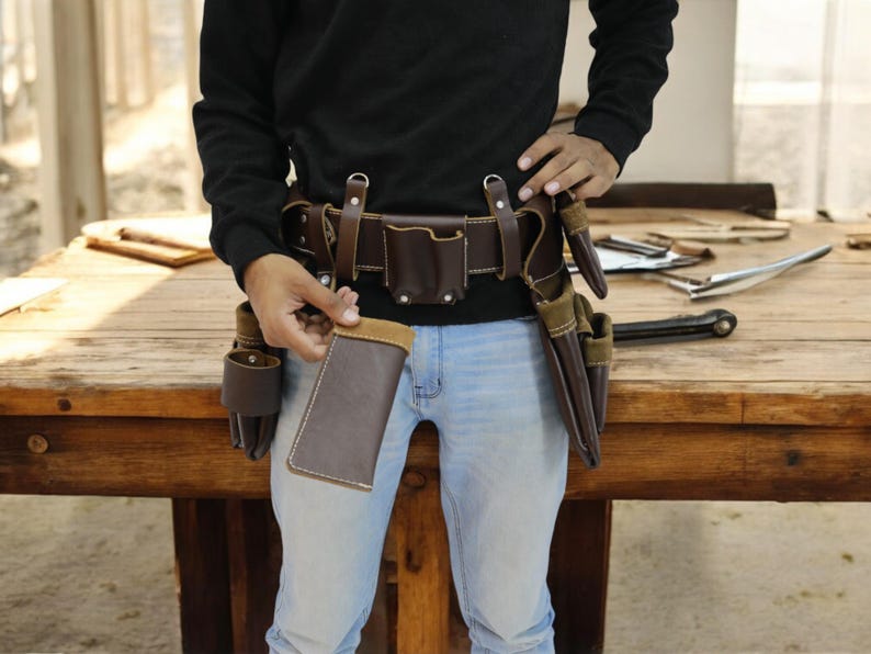 May include: A brown leather tool belt with multiple pockets and pouches. The belt is worn by a person and is secured with a metal buckle. The belt is on a wooden surface.