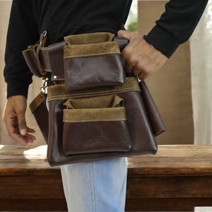 May include: A brown leather tool belt with multiple pockets, including a suede pocket. The belt is worn by a person and is secured with a metal buckle.