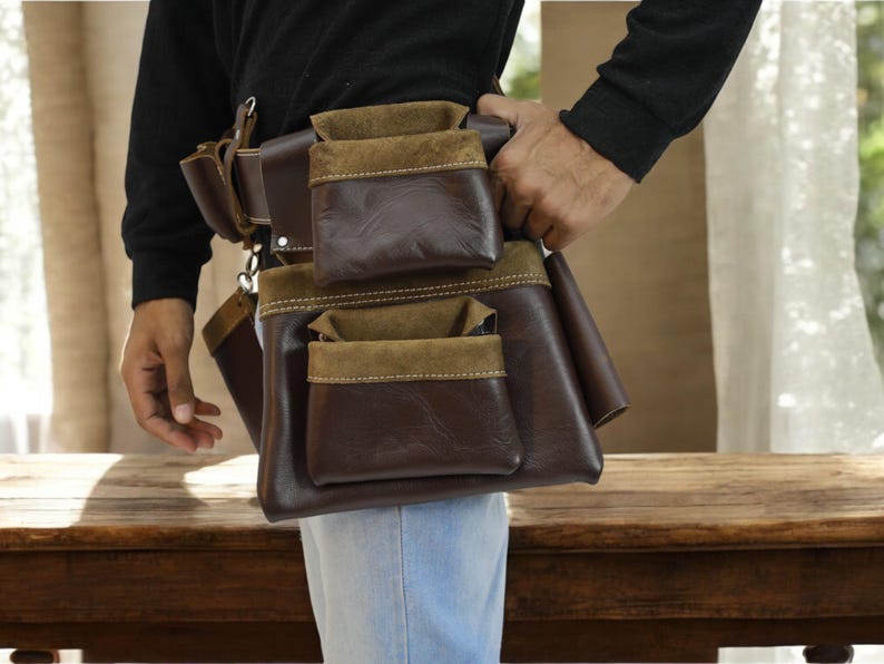 May include: A brown leather tool belt with multiple pockets, including a suede pocket. The belt is worn by a person and is secured with a metal buckle.