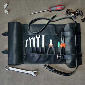 Puede incluir: Rollo de herramientas de cuero negro con varias herramientas, incluyendo llaves, alicates y un destornillador. El rollo se asegura con correas y hebillas. También hay una pequeña escultura de motocicleta de metal y dos objetos plateados.