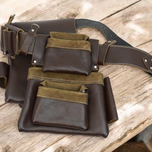 May include: A brown leather tool belt with multiple pockets and a brown leather belt with a silver buckle.