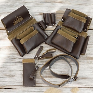 Può includere: Un set di cinture portautensili in pelle marrone con più tasche e una cinghia in pelle marrone. Le cinture sono su una superficie in legno chiaro.