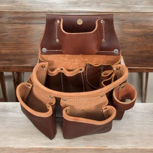 May include: A brown leather tool belt with multiple pockets and compartments. The belt features a variety of sizes and shapes, including a hammer loop and several pouches. The interior is a lighter tan color, and the stitching is white.