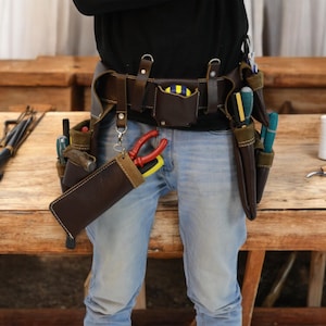May include: A brown leather tool belt with multiple pockets and pouches, worn over blue jeans. The belt is filled with various tools, including screwdrivers, pliers, and a hammer.