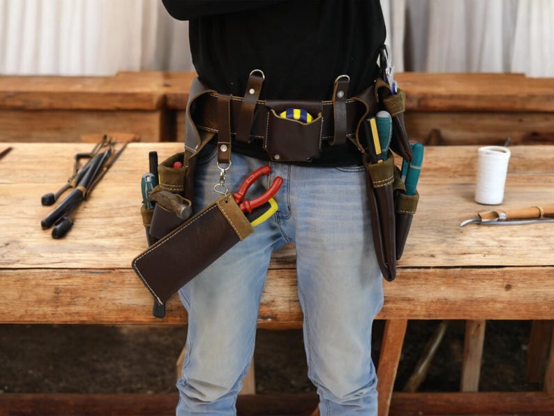 May include: A brown leather tool belt with multiple pockets and pouches, worn over blue jeans. The belt is filled with various tools, including screwdrivers, pliers, and a hammer.