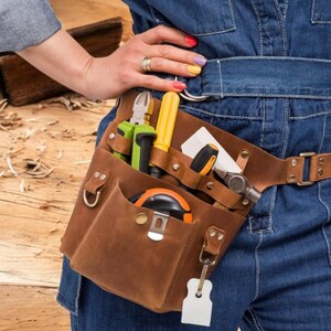 Pu&ograve; includere: Una cintura portautensili in pelle marrone con pi&ugrave; tasche e una variet&agrave; di strumenti, tra cui un martello, un metro a nastro e cacciaviti. La cintura &egrave; indossata da una persona che indossa jeans blu.