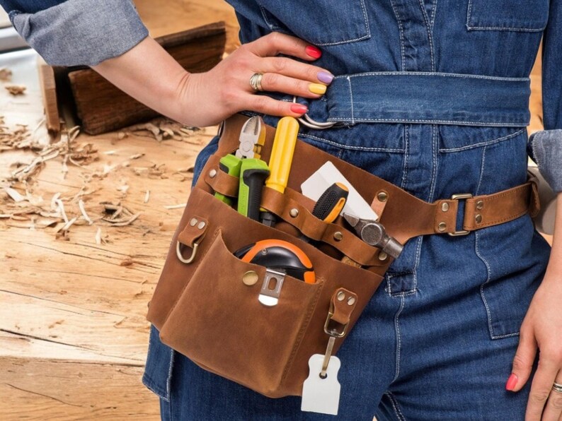 Pu&ograve; includere: Una cintura portautensili in pelle marrone con pi&ugrave; tasche e una variet&agrave; di strumenti, tra cui un martello, un metro a nastro e cacciaviti. La cintura &egrave; indossata da una persona che indossa jeans blu.