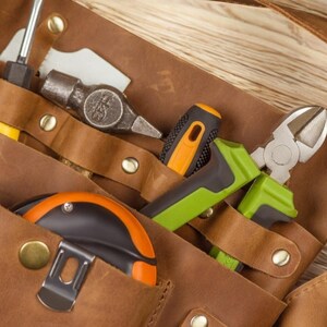 Pu&ograve; includere: Una cintura portautensili in pelle marrone con vari strumenti, tra cui un martello, una pinza, un cacciavite e un metro a nastro.