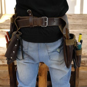 May include: A brown leather tool belt with multiple pockets and pouches, worn over blue jeans. The belt has a metal buckle and is secured with a brown leather strap.